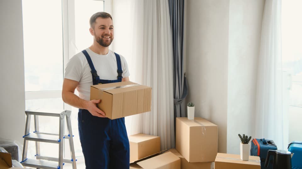 A smiling mover in blue overalls carrying a packed moving box during pickup from an apartment by DACHSER & KOLB.