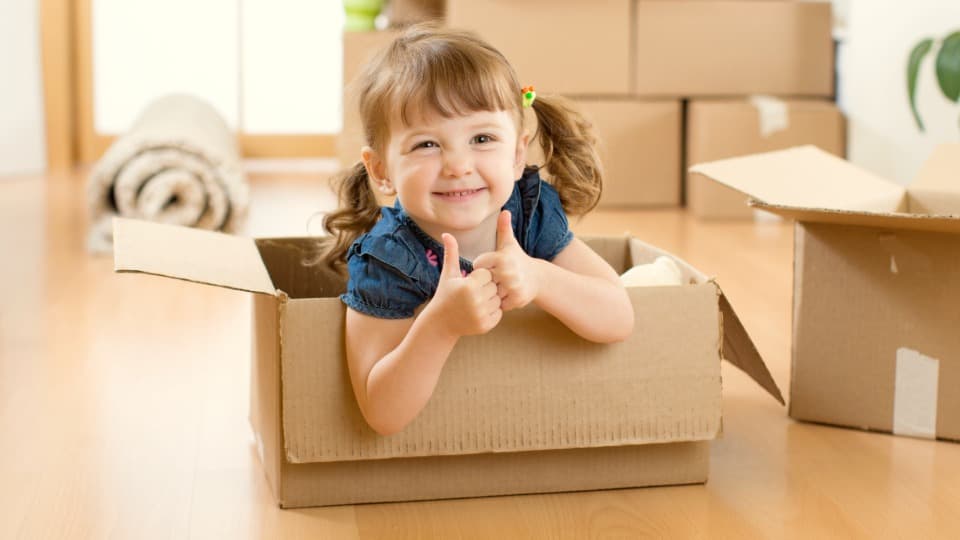 Ein fröhliches kleines Mädchen zeigt in einem Umzugskarton beide Daumen nach oben für einen entspannten Auslandsumzug mit Kindern mit DACHSER & KOLB.