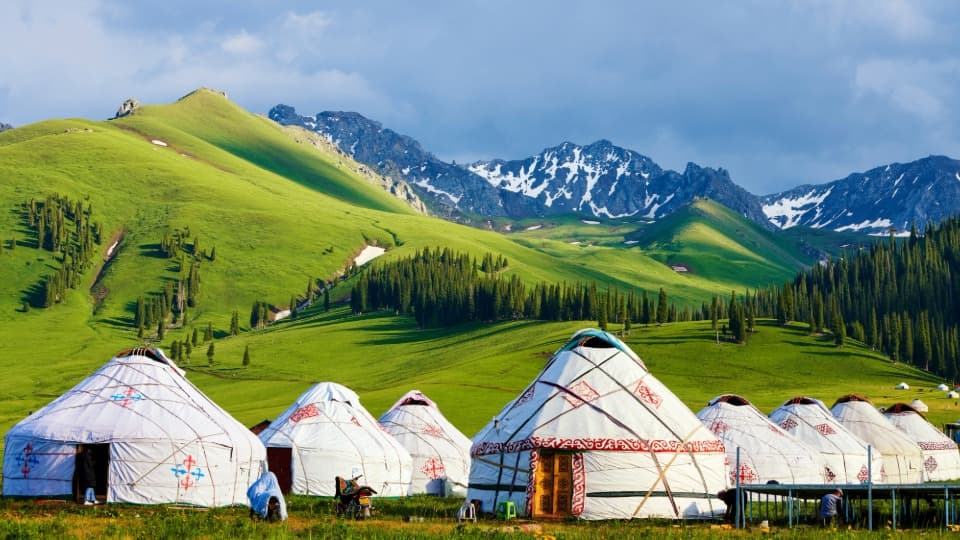 Traditionelle Jurten in grüner Berglandschaft der Mongolei mit schneebedeckten Gipfeln im Hintergrund