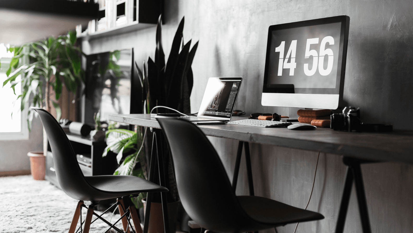 Graues Home Office nach Umzug mit langem Schreibtisch, zwei Stühlen, Laptop, Monitor mit Uhrzeit und Pflanzen. Einrichtungsideen für gemeinsames Arbeiten.