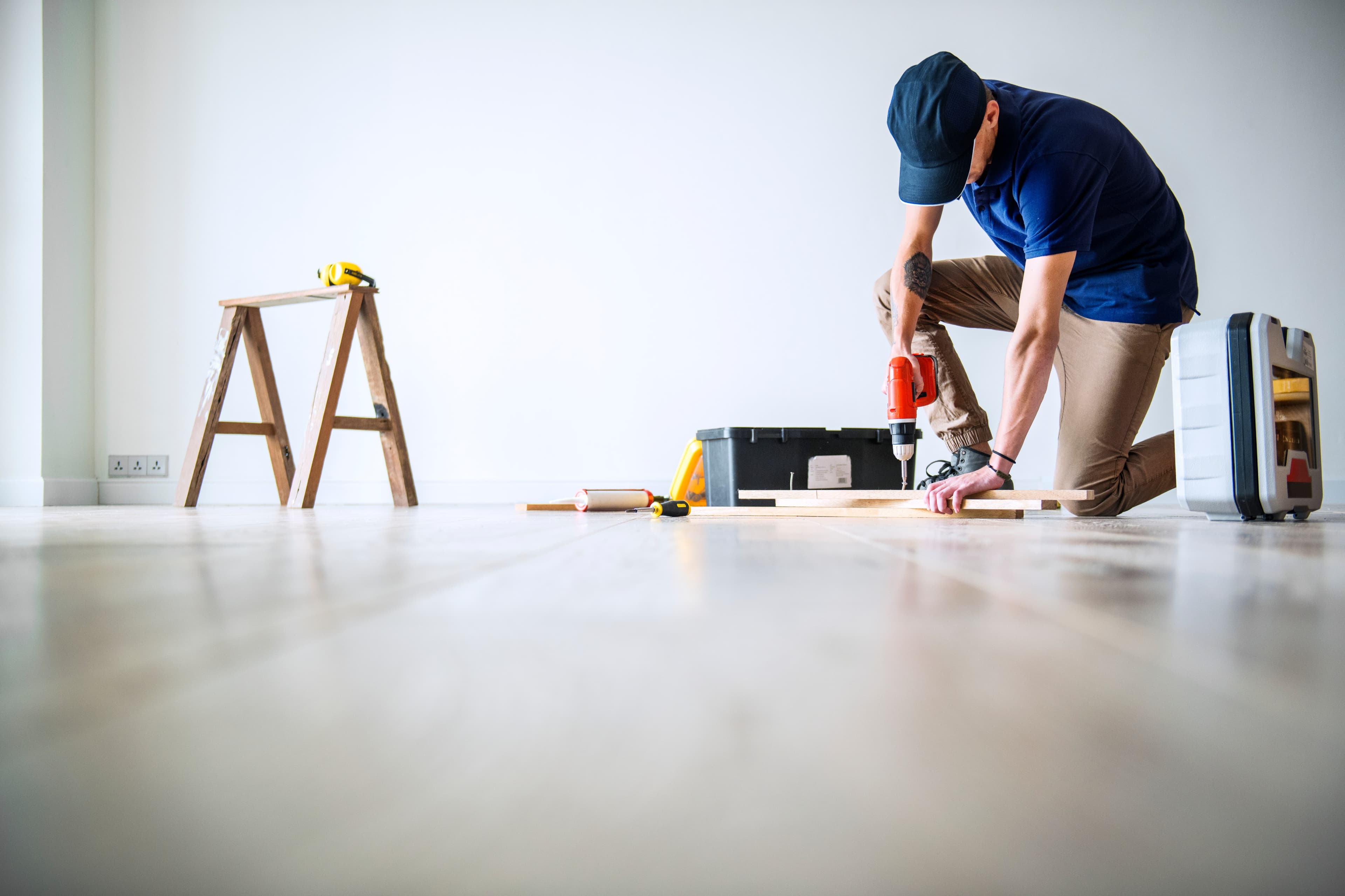 Handwerkerservice: Mann kniet auf Holzboden, bohrt Loch, Werkzeugkasten und Leiter im Hintergrund.