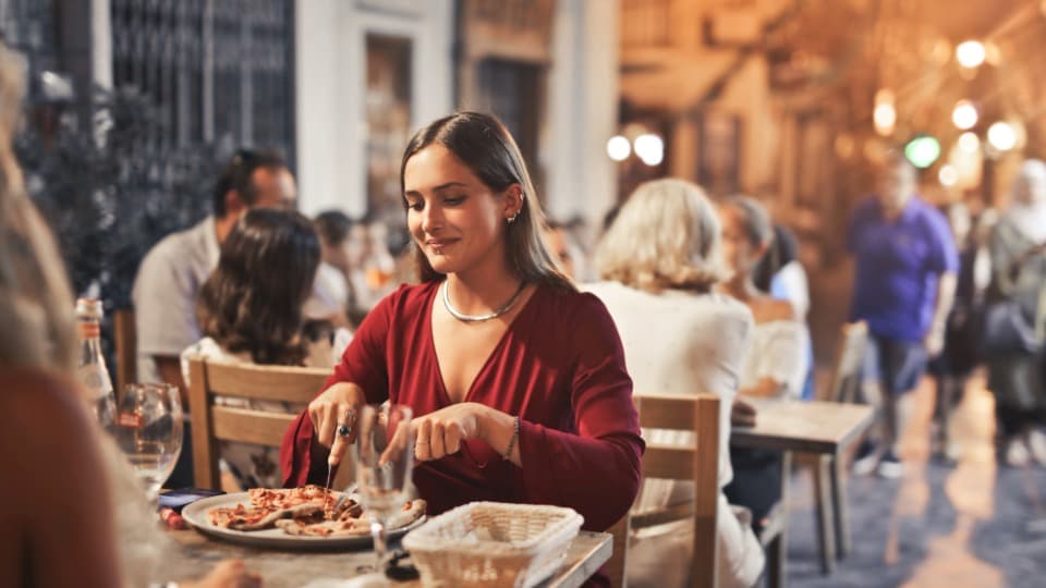 Eine lächelnde Frau isst abends Pizza in einem belebten Restaurant im Freien – typisch für das mediterrane Lebensgefühl und die Kultur Maltas.