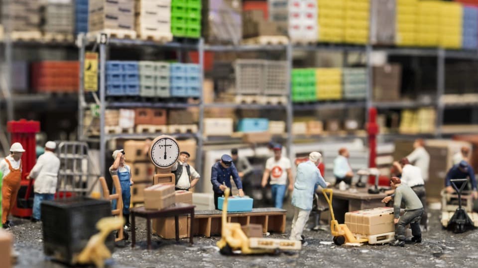 Miniatur-Figuren auf einer Modelleisenbahnanlage beim Verpacken und Wiegen von Frachtgut als Symbol für die Umzugsplanung durch DACHSER & KOLB.