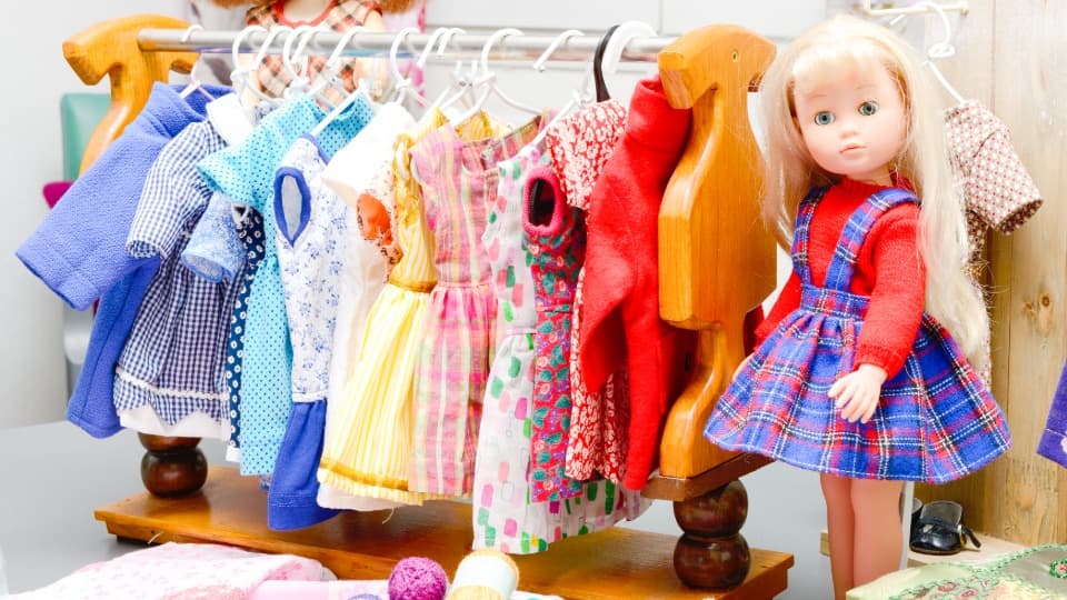 Eine blonde Sammlerpuppe steht neben einer Miniatur-Kleiderstange mit Puppenkleidung vor dem Schutz des Zubehörs für den Umzug mit DACHSER & KOLB.