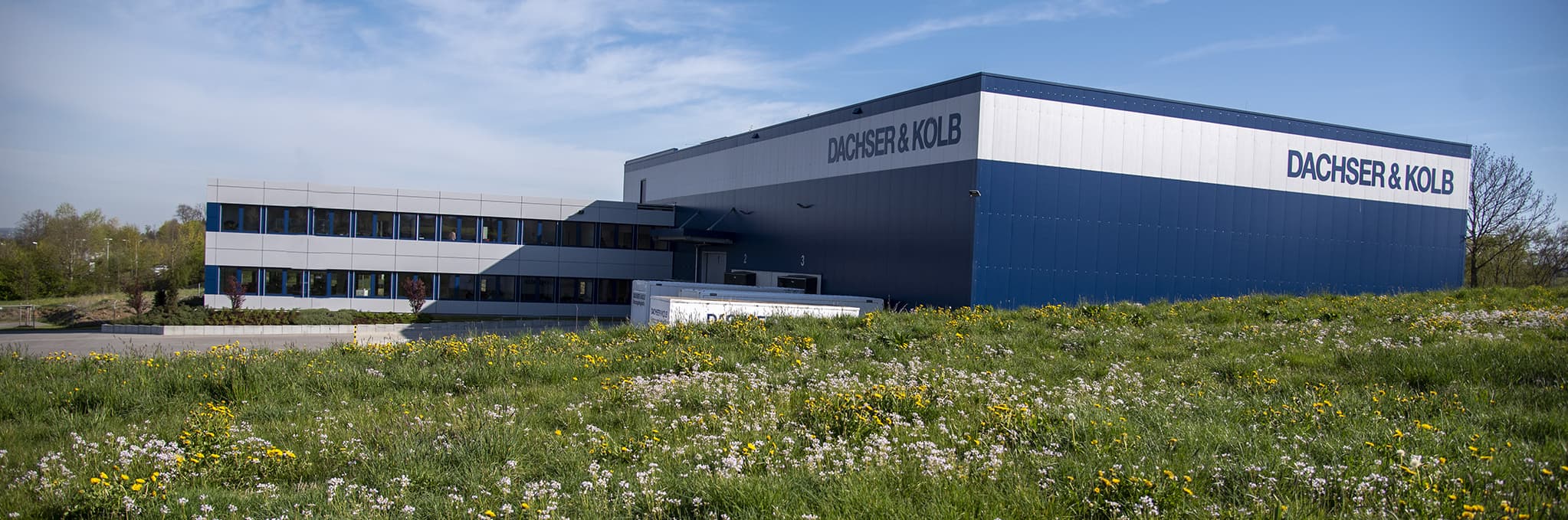 Modernes Gebäude des Umzugsunternehmens DACHSER & KOLB mit Lagerhalle und Bürotrakt, umgeben von grüner Wiese.