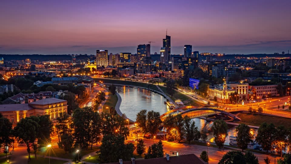 Nächtliche Skyline von Vilnius mit beleuchteten Hochhäusern am Fluss Neris – Symbol für die Stabilität und Sicherheit im modernen Litauen.