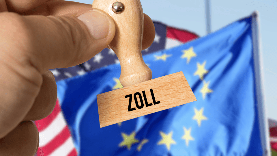 Hand hält Holzstempel mit Aufschrift ZOLL vor Flaggen der USA und EU.